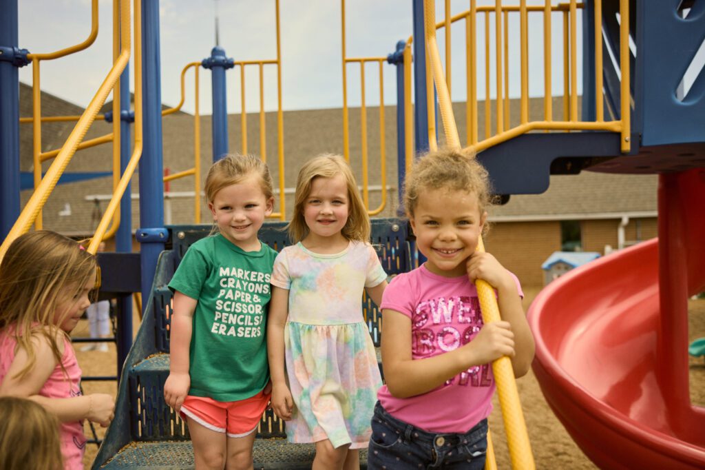 Three Preschool students smile at the camera from the playground steps..