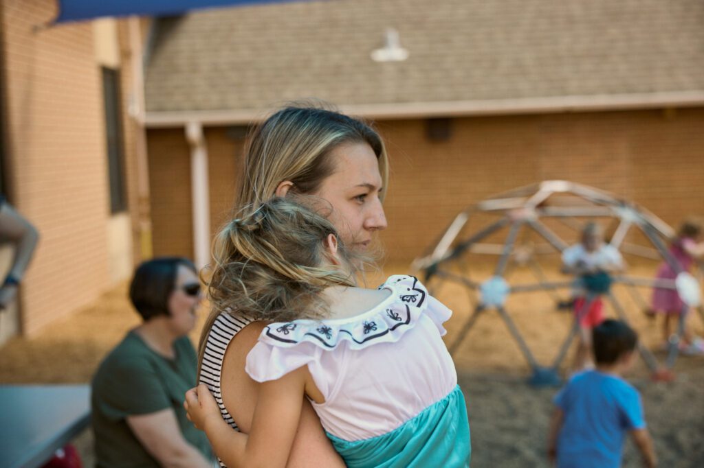 Teacher holds preschool student while watching over the rest of the class play on playground.