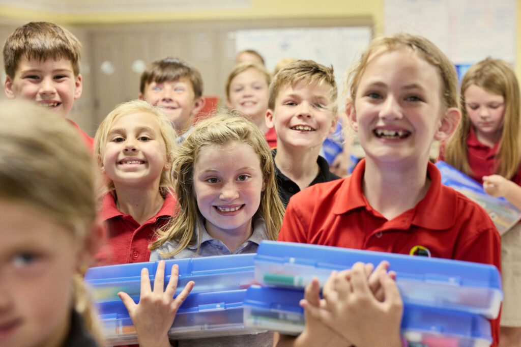 Elementary class smiles excitedly at the camera holding up school supplies.