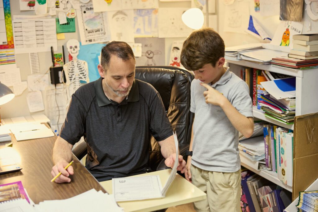 A male teacher talks over a boy's work with him at desk.