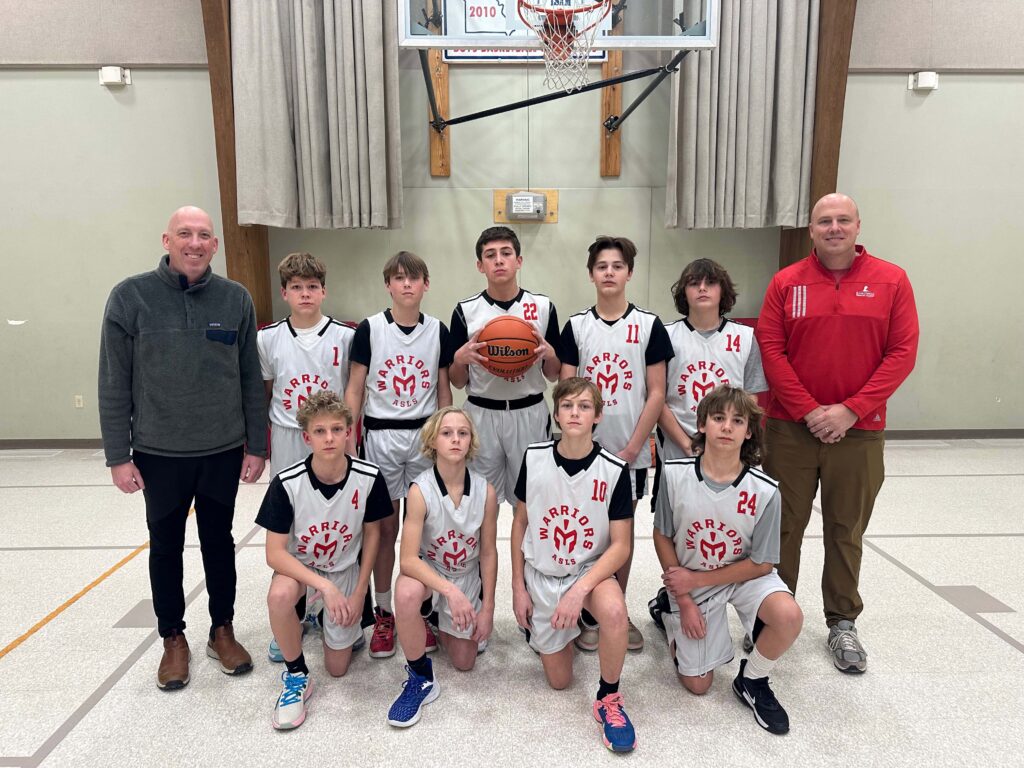 Abiding Savior Lutheran School Boys Basketball takes a picture with their coach on the court in front of the basketball hoop.